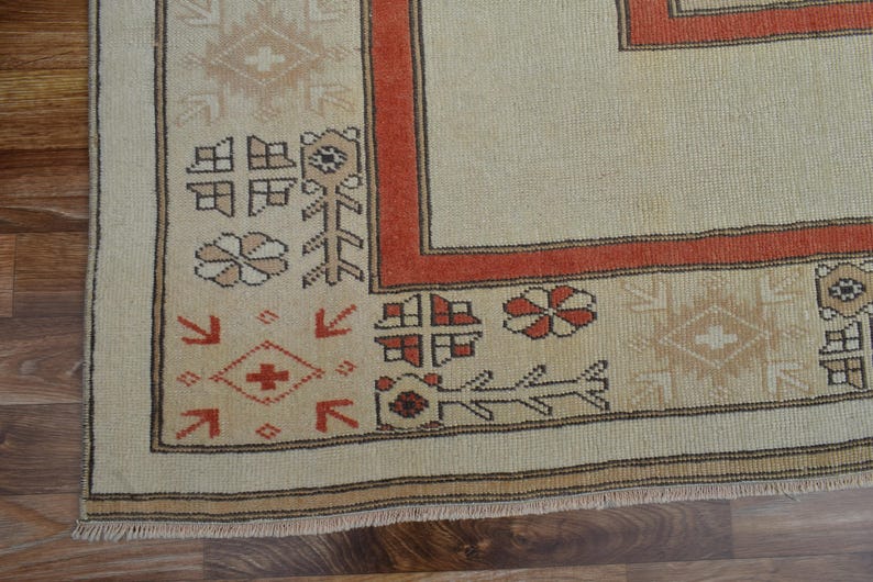May include: Close-up of a beige and rust-colored rug with intricate geometric patterns. The rug features a border with repeating floral and geometric motifs in shades of rust, beige, and brown. The rug's design is a traditional style.