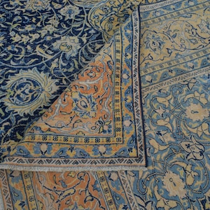 K&ouml;nnte beinhalten: Ein aufwendig gemusterter Teppich mit einer gefalteten Ecke, der eine reiche Farbpalette zeigt. Die dominierenden Farbt&ouml;ne sind tiefes Marineblau, Hellblau und ein warmes Pfirsich. Blumen- und Arabeskenmuster schaffen eine klassische, elegante &Auml;sthetik.