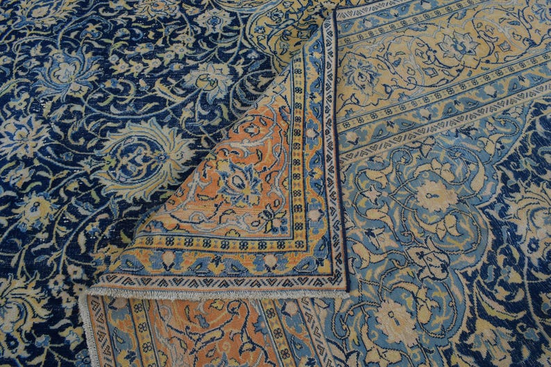 K&ouml;nnte beinhalten: Ein aufwendig gemusterter Teppich mit einer gefalteten Ecke, der eine reiche Farbpalette zeigt. Die dominierenden Farbt&ouml;ne sind tiefes Marineblau, Hellblau und ein warmes Pfirsich. Blumen- und Arabeskenmuster schaffen eine klassische, elegante &Auml;sthetik.