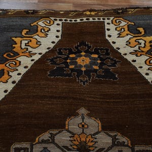 May include: A close-up of a brown, grey, and cream rug with an intricate geometric design. The rug features a central medallion and decorative borders with floral and scroll patterns. The rug's dimensions are not visible.