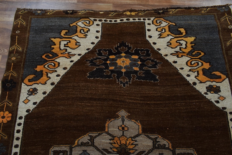 May include: A close-up of a brown, grey, and cream rug with an intricate geometric design. The rug features a central medallion and decorative borders with floral and scroll patterns. The rug's dimensions are not visible.
