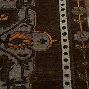 May include: Close-up of a section of a brown and grey patterned rug. The rug features floral and geometric designs in shades of brown, grey, and orange. A cream-coloured border with black dots and floral accents runs along the edge.