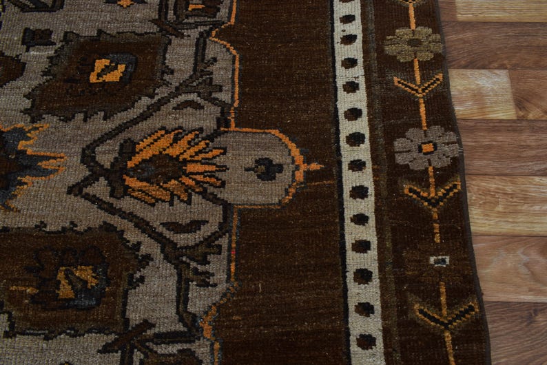 May include: Close-up of a section of a brown and grey patterned rug. The rug features floral and geometric designs in shades of brown, grey, and orange. A cream-coloured border with black dots and floral accents runs along the edge.