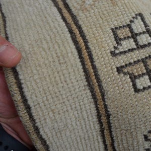 May include: Close-up of a beige and brown patterned rug. The rug has a textured surface with geometric shapes and vertical stripes. The edge of the rug is visible, held by a hand.