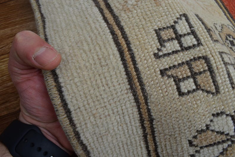 May include: Close-up of a beige and brown patterned rug. The rug has a textured surface with geometric shapes and vertical stripes. The edge of the rug is visible, held by a hand.