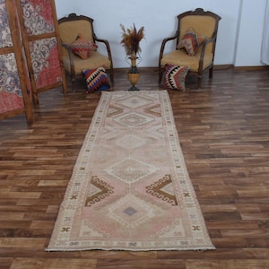 May include: A long, rectangular rug with a faded, vintage appearance. The rug features a central diamond pattern in shades of beige, pink, and brown. The rug is placed on a wooden floor, with two chairs and decorative pillows in the background.