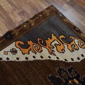 May include: A section of a brown, grey, and orange patterned rug. The rug features a dark brown field, a grey border, and a cream-coloured section with orange and black decorative elements. The rug is on a wooden floor.