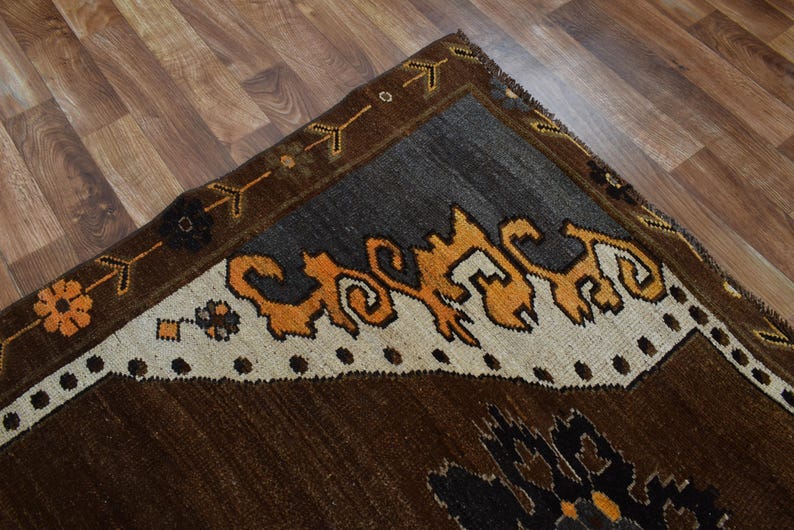 May include: A section of a brown, grey, and orange patterned rug. The rug features a dark brown field, a grey border, and a cream-coloured section with orange and black decorative elements. The rug is on a wooden floor.