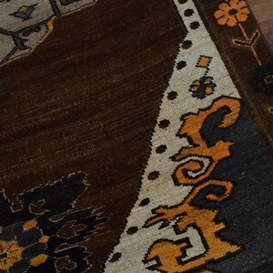 May include: Detail of a patterned rug in shades of brown, beige, orange, black, and grey. The rug's design incorporates floral and geometric patterns. The border is beige with orange floral accents, and the field is dark brown.
