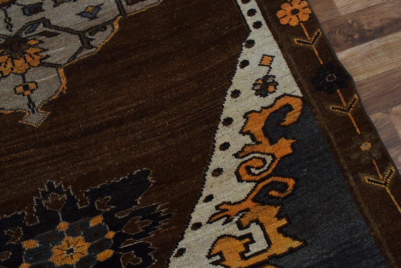 May include: Detail of a patterned rug in shades of brown, beige, orange, black, and grey. The rug's design incorporates floral and geometric patterns. The border is beige with orange floral accents, and the field is dark brown.