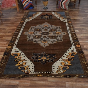 May include: A rectangular brown rug with a central medallion design in shades of grey, beige, and yellow. The rug has a dark brown field and a border with floral and geometric patterns. The rug is placed on a wooden floor.