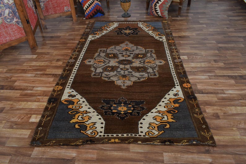 May include: A rectangular brown rug with a central medallion design in shades of grey, beige, and yellow. The rug has a dark brown field and a border with floral and geometric patterns. The rug is placed on a wooden floor.