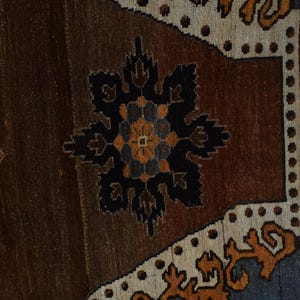 May include: Close-up of a section of a brown and beige rug with a central black and orange floral design. The rug has a textured appearance with a border of beige and blue geometric patterns. The rug is likely handmade.