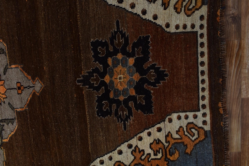 May include: Close-up of a section of a brown and beige rug with a central black and orange floral design. The rug has a textured appearance with a border of beige and blue geometric patterns. The rug is likely handmade.
