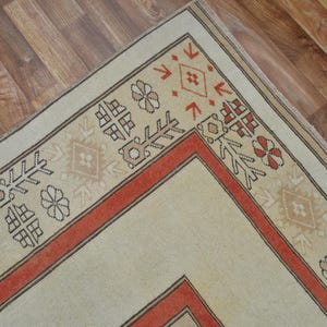 May include: A section of a beige and red patterned rug. The rug features a central beige field with a red border and decorative geometric and floral motifs in brown and red. The rug is placed on a wooden floor.