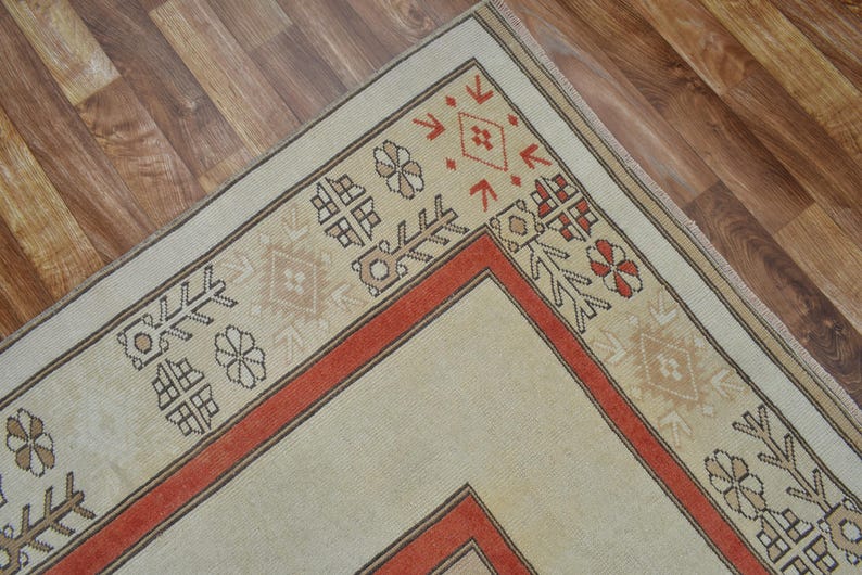 May include: A section of a beige and red patterned rug. The rug features a central beige field with a red border and decorative geometric and floral motifs in brown and red. The rug is placed on a wooden floor.