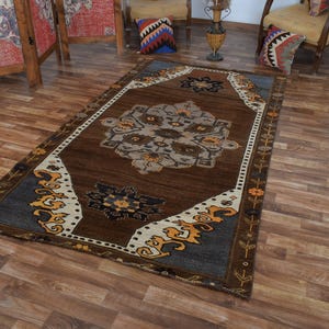 May include: A rectangular brown rug with a central medallion design in grey, beige, and blue. The rug has a dark brown border with decorative floral and geometric patterns in yellow and white. The rug is on a wooden floor.