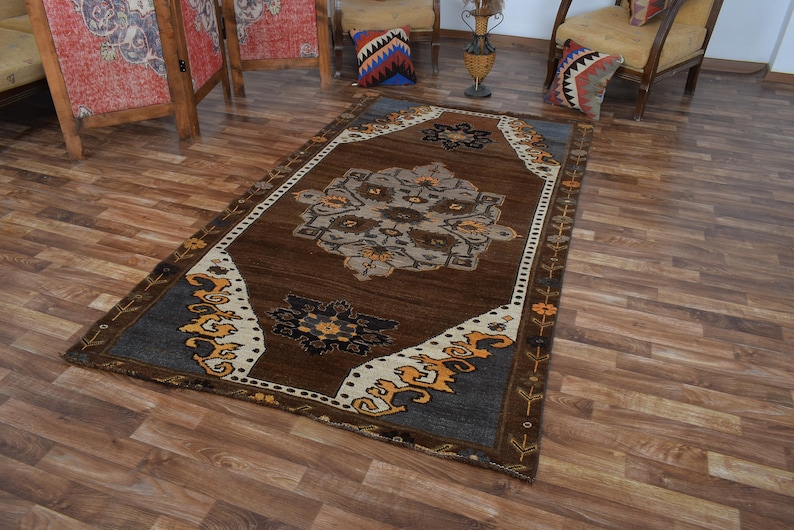 May include: A rectangular brown rug with a central medallion design in grey, beige, and blue. The rug has a dark brown border with decorative floral and geometric patterns in yellow and white. The rug is on a wooden floor.