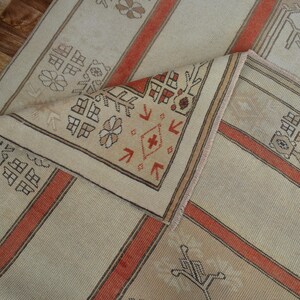 May include: Beige and orange patterned rug with a folded corner. The rug features a repeating geometric design with floral and linear elements. The color scheme includes beige, orange, and brown, creating a warm and inviting aesthetic. The rug is likely made of woven fabric.