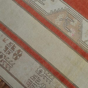 May include: Close-up of a section of a vintage rug with a cream-colored field and rust-colored stripes. The rug features geometric patterns in shades of brown and beige, creating a warm and inviting aesthetic. The rug is likely made of wool.