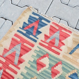 May include: Close-up of a colorful woven rug with geometric patterns in shades of red, blue, green, and beige. The rug has a fringed edge and is made of a soft, textured material.