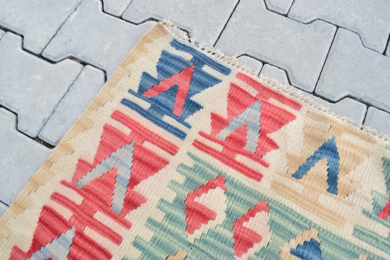May include: Close-up of a colorful woven rug with geometric patterns in shades of red, blue, green, and beige. The rug has a fringed edge and is made of a soft, textured material.