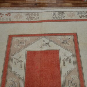 May include: A rectangular, vintage rug with a central red panel framed by a beige border and a rust-colored border. The rug features geometric patterns in beige, rust, and brown, creating a traditional aesthetic. The rug is placed on a wooden floor.