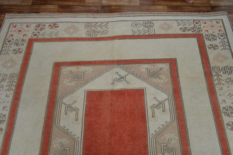 May include: A rectangular, vintage rug with a central red panel framed by a beige border and a rust-colored border. The rug features geometric patterns in beige, rust, and brown, creating a traditional aesthetic. The rug is placed on a wooden floor.