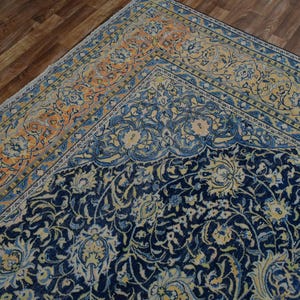 K&ouml;nnte beinhalten: Ausschnitt eines verzierten Vintage-Teppichs mit marineblauem Feld und einem Rand in Orange- und Blaut&ouml;nen. Der Teppich weist ein detailliertes Blumenmuster auf und schafft eine klassische und elegante &Auml;sthetik. Der Teppich liegt auf einem Holzboden.