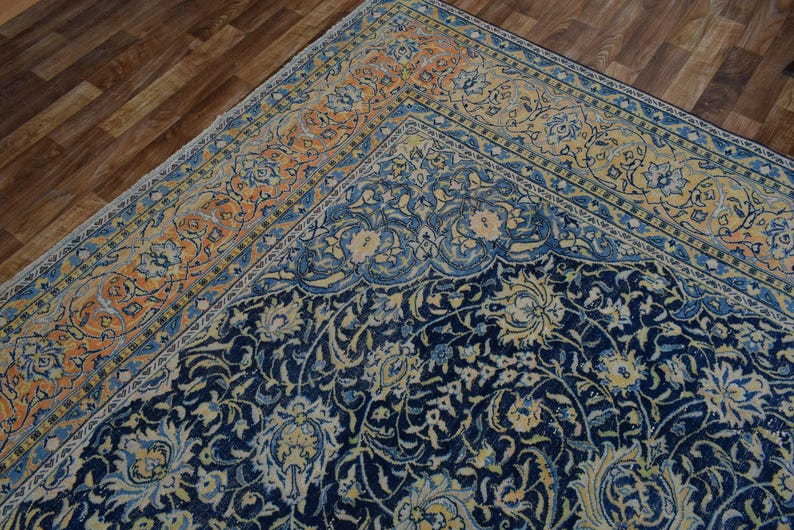 K&ouml;nnte beinhalten: Ausschnitt eines verzierten Vintage-Teppichs mit marineblauem Feld und einem Rand in Orange- und Blaut&ouml;nen. Der Teppich weist ein detailliertes Blumenmuster auf und schafft eine klassische und elegante &Auml;sthetik. Der Teppich liegt auf einem Holzboden.