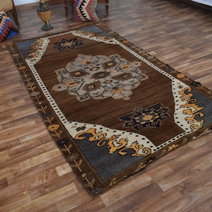 May include: A rectangular brown rug with a central floral design in shades of grey, beige, and brown. The rug has a dark blue border with orange and white accents. The rug is placed on a wooden floor.
