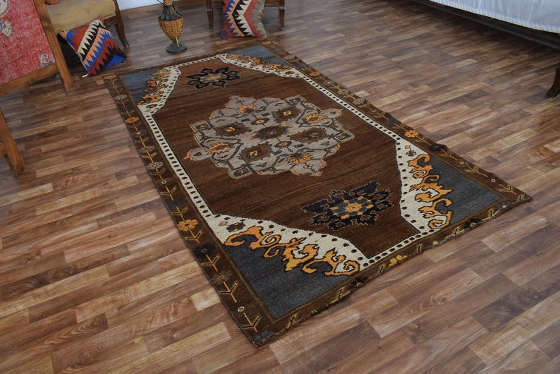 May include: A rectangular brown rug with a central floral design in shades of grey, beige, and brown. The rug has a dark blue border with orange and white accents. The rug is placed on a wooden floor.