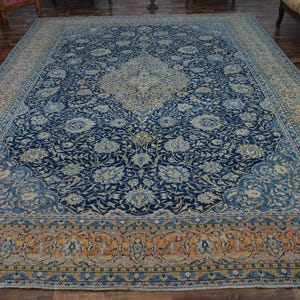K&ouml;nnte beinhalten: Gro&szlig;er, rechteckiger Teppich mit einem komplizierten Blumenmuster in Marineblau, Creme und Orange. Der Teppich hat ein zentrales Medaillon-Design und einen detaillierten Rand. Der Teppich liegt auf einem Holzboden.