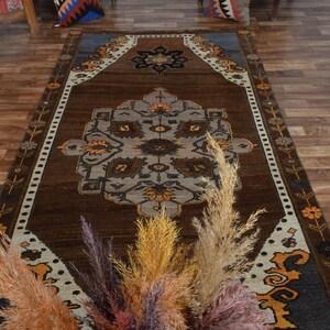 May include: A brown rectangular rug with a central medallion design in grey, orange, and black. The rug has a border with floral and geometric patterns. Dried ornamental grasses in various colours are in the foreground.