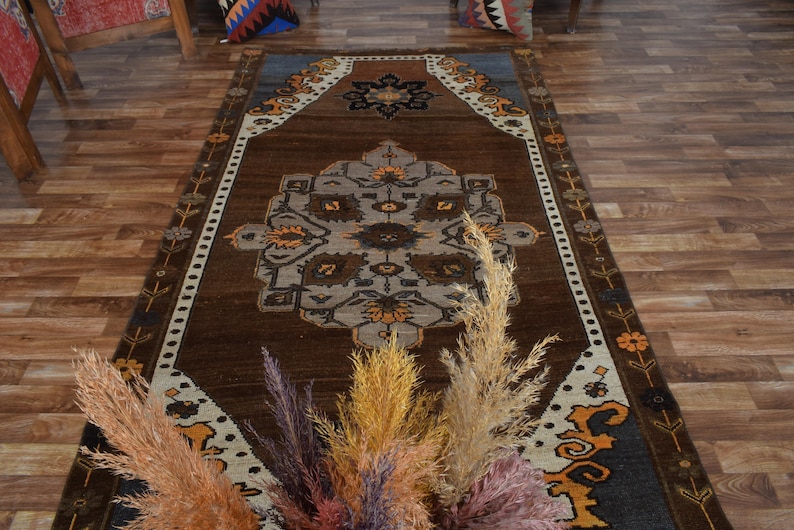 May include: A brown rectangular rug with a central medallion design in grey, orange, and black. The rug has a border with floral and geometric patterns. Dried ornamental grasses in various colours are in the foreground.
