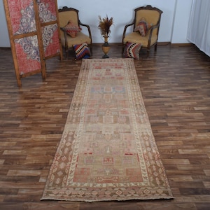 Könnte beinhalten: Ein rechteckiger, alter Teppich mit einem verblassten, abgenutzten Aussehen. Der Teppich hat ein zentrales geometrisches Muster in Beige-, Rot- und Blautönen mit einem braunen Rand. Er liegt auf einem Holzboden.