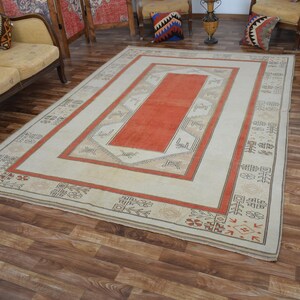 May include: Rectangular area rug with a central orange panel, framed by a cream border and a red outer border. The rug features geometric patterns and decorative elements, suitable for living room decor.