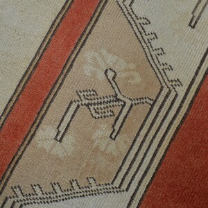 May include: Close-up of a section of a rug with a geometric design. The rug features a beige background with a diagonal stripe in burnt orange and a central panel with a repeating pattern in beige and black.