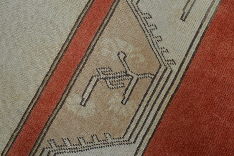 May include: Close-up of a section of a rug with a geometric design. The rug features a beige background with a diagonal stripe in burnt orange and a central panel with a repeating pattern in beige and black.
