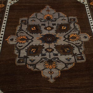 May include: A rectangular brown rug featuring a central geometric pattern in grey, brown, and orange. The rug has a dark brown border and floral accents along the edges. It is placed on a wooden floor.