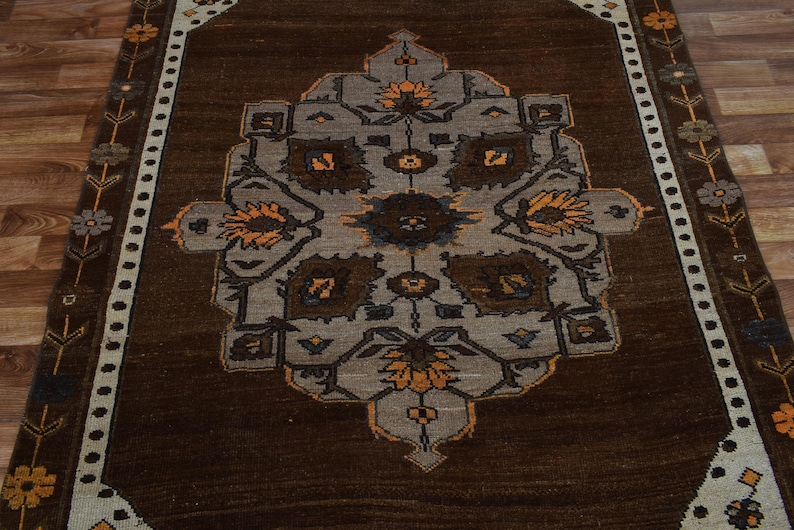 May include: A rectangular brown rug featuring a central geometric pattern in grey, brown, and orange. The rug has a dark brown border and floral accents along the edges. It is placed on a wooden floor.