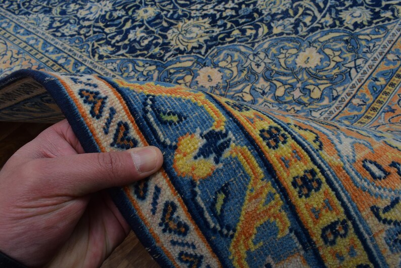 K&ouml;nnte beinhalten: Ausschnitt eines detaillierten, verzierten Teppichs mit umgeschlagenem Rand. Der Teppich weist ein komplexes Blumenmuster in den Farbt&ouml;nen Marineblau, Hellblau, Gelb und Orange auf. Der umgeschlagene Rand zeigt den mehrschichtigen Aufbau und das komplizierte Design des Teppichs.