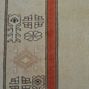 May include: Close-up of a beige rug with a geometric pattern and a red border. The rug features repeating shapes and floral motifs in brown and beige tones, creating a traditional design.