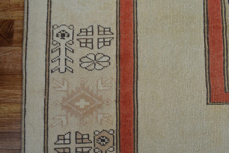 May include: Close-up of a beige rug with a geometric pattern and a red border. The rug features repeating shapes and floral motifs in brown and beige tones, creating a traditional design.