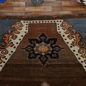 May include: A rectangular brown rug with a central floral design in black, blue, and orange. The rug features a border with stylized orange and white patterns against a blue and brown background. The rug is made of woven material.