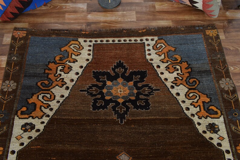 May include: A rectangular brown rug with a central floral design in black, blue, and orange. The rug features a border with stylized orange and white patterns against a blue and brown background. The rug is made of woven material.