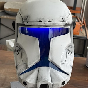 May include: A white tactical helmet with a weathered, distressed finish and black scuff marks. The helmet features a prominent T-shaped visor with a vibrant, glowing blue translucent lens, integrated side vents, and a central chin ventilation port.