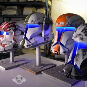 May include: Four replica clone trooper helmets displayed on stands. The first has red claw marks, the second is white with green accents, the third is orange and white, and the fourth is black and white. Each helmet features a glowing blue visor.