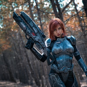 Peut inclure: Une personne porte un costume futuriste blindé et tient une grosse arme. Le costume est principalement gris et noir avec des détails rouges. L'arrière-plan est une scène de forêt floue avec des couleurs automnales.