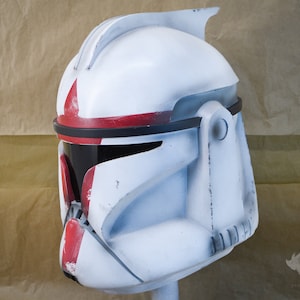 May include: A white and red Star Wars clone trooper helmet with a black visor. The helmet has a weathered look and is made of plastic.